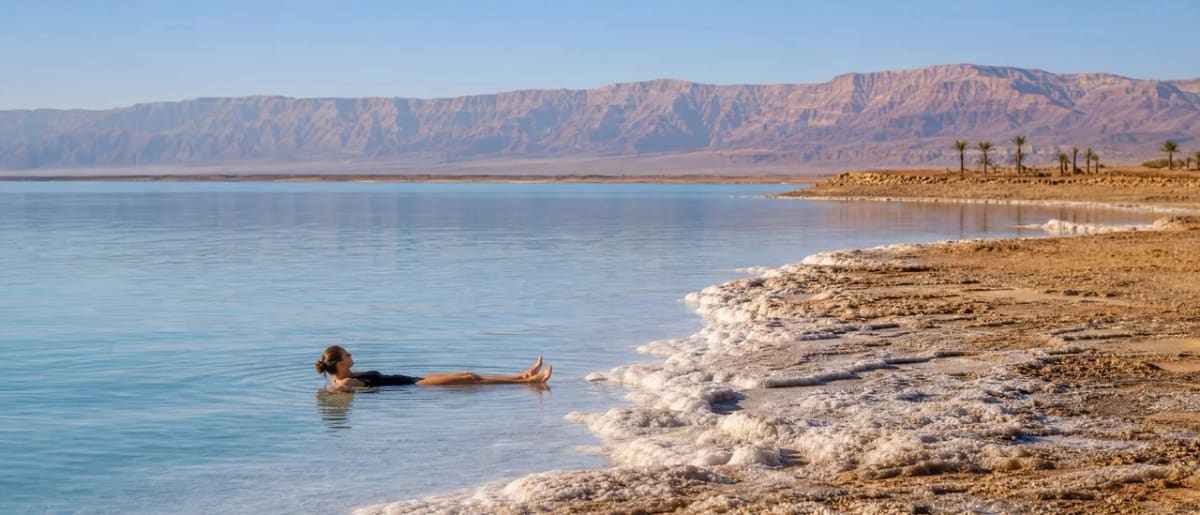 A traveler floating effortlessly in the mineral-rich blue water of the Dead Sea with mountains in the background